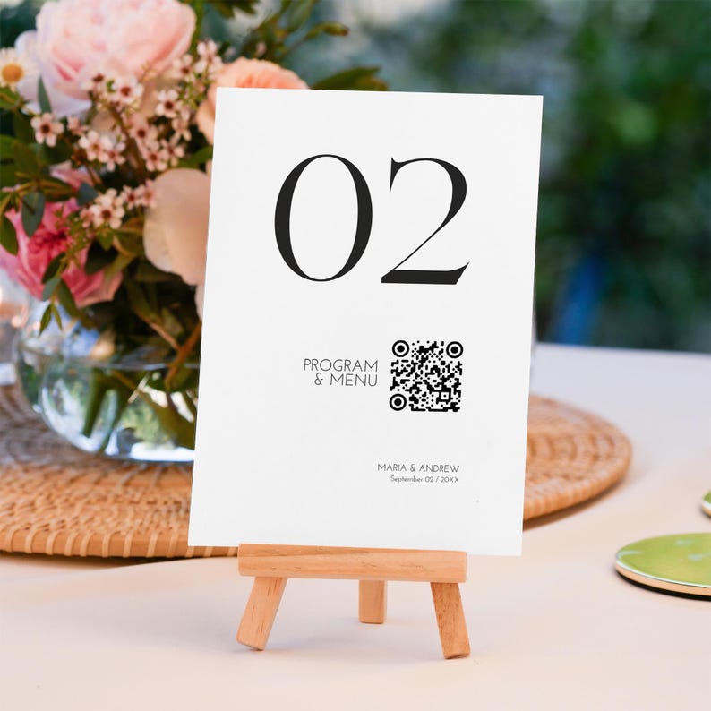 Op de afbeelding: Een witte tafelnummerkaart met het nummer "02" in grote zwarte letters. De kaart bevat ook de tekst "PROGRAMMA & MENU", een QR-code en namen met een datum. De kaart wordt weergegeven op een kleine houten ezel.