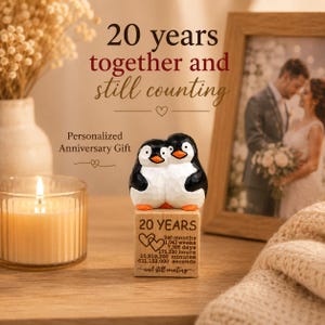 May include: A wooden anniversary gift featuring two carved penguins embracing. The penguins sit atop a wooden block engraved with "20 YEARS" and time measurements. The image includes a lit candle, a framed photo, and text that reads "20 years together and still counting."