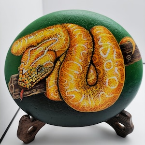 May include: A hand-painted round rock with a green background and a yellow snake with white markings coiled around a branch. The rock is on a wooden stand.