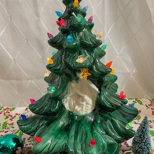 May include: A green ceramic Christmas tree with a white ceramic nativity scene inside. The tree is decorated with colourful lights and a gold star on top.