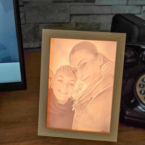 Könnte beinhalten: Ein gerahmtes Foto mit einem warm getönten Bild einer Frau und eines Kindes. Der Holzrahmen umgibt das Porträt, das mit einem sanften, orangefarbenen Schein beleuchtet wird. Die Personen lächeln und schaffen eine herzerwärmende Szene.