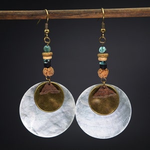 Könnte beinhalten: Ein Paar Hängeohrringe mit einer großen weißen Muschelscheibe, einer Messing-Scheibe und braunen Holzperlen. Die Perlen sind türkis, braun, schwarz und beige.