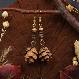 May include: A pair of gold tone earrings with brown and beige wooden beads and pine cone charms. The earrings are displayed on a brown leaf with other autumn foliage.