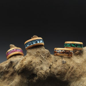 Puede incluir: Cuatro anillos de madera con diseños de incrustaciones coloridas. Los anillos están dispuestos sobre un trozo de madera a la deriva.