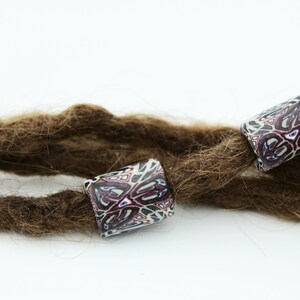 May include: Three brown dreadlocks with colorful patterned beads. The beads are cylindrical and have a white background with pink, purple, and black geometric designs.