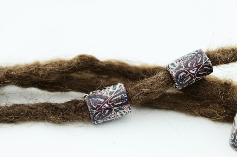 May include: Three brown dreadlocks with colorful patterned beads. The beads are cylindrical and have a white background with pink, purple, and black geometric designs.