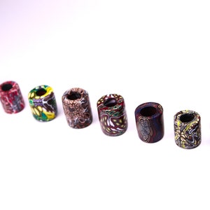 May include: A set of eight colorful, patterned, cylindrical beads. The beads are made of a hard, durable material and have a smooth, polished finish. The beads are arranged in a row on a white background.