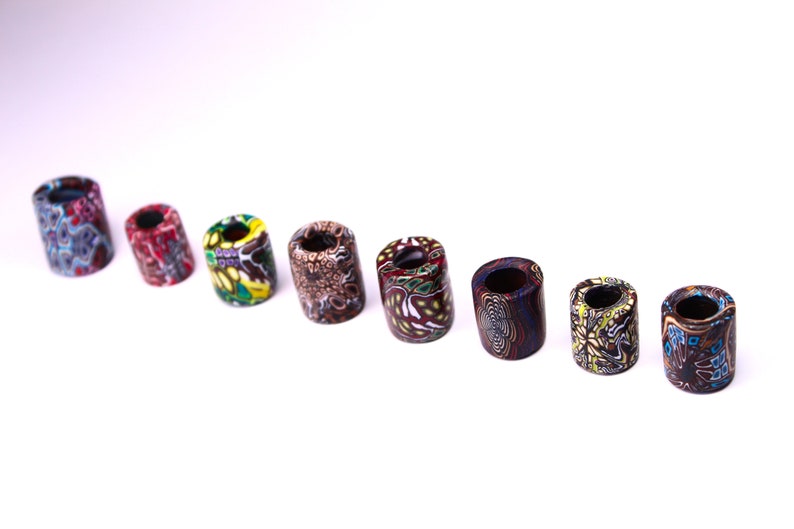 May include: A set of eight colorful, patterned, cylindrical beads. The beads are made of a hard, durable material and have a smooth, polished finish. The beads are arranged in a row on a white background.