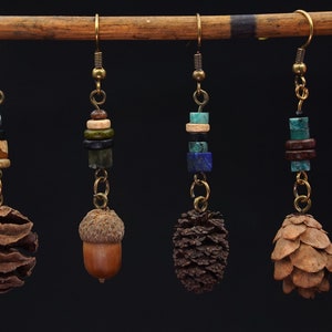 Puede incluir: Cuatro pares de pendientes colgantes con un tema de naturaleza. Cada pendiente presenta un elemento natural diferente, incluyendo una piña, una bellota y una variedad de cuentas de piedras preciosas en tonos azules, verdes y marrones.