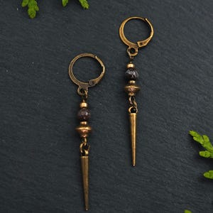 Puede incluir: Un par de pendientes de color oro antiguo. Cada pendiente presenta un cierre de palanca, una serie de cuentas de color marrón oscuro y una espiga larga y puntiaguda. Los pendientes se exhiben sobre una superficie gris oscuro con vegetación y una piedra.