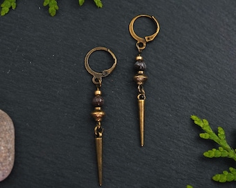 Può includere: Un paio di orecchini color oro antico. Ogni orecchino presenta una chiusura a leva, una serie di perline marrone scuro e una lunga punta. Gli orecchini sono esposti su una superficie grigio scuro con vegetazione e una pietra.