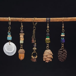 Könnte beinhalten: Fünf Paar Ohrringe mit naturinspirierten Designs. Jeder Ohrring besteht aus einer anderen Kombination aus Holz-, Stein- und Metallperlen. Die Ohrringe hängen an einer Holzstange.