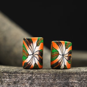 May include: Two colorful cylindrical beads with a white, orange, green, and brown floral pattern. The beads are sitting on a rough, gray surface.