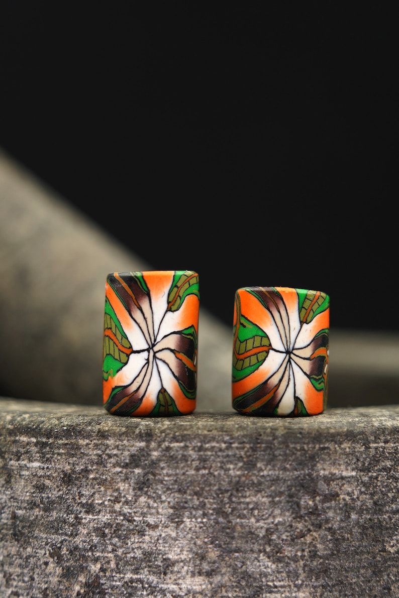 May include: Two colorful cylindrical beads with a white, orange, green, and brown floral pattern. The beads are sitting on a rough, gray surface.