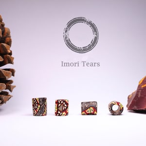 May include: Four colorful glass beads with a geometric pattern in shades of brown, red, and yellow. The beads are arranged in a row on a white surface. The text "I mori Tears" is visible in the background.