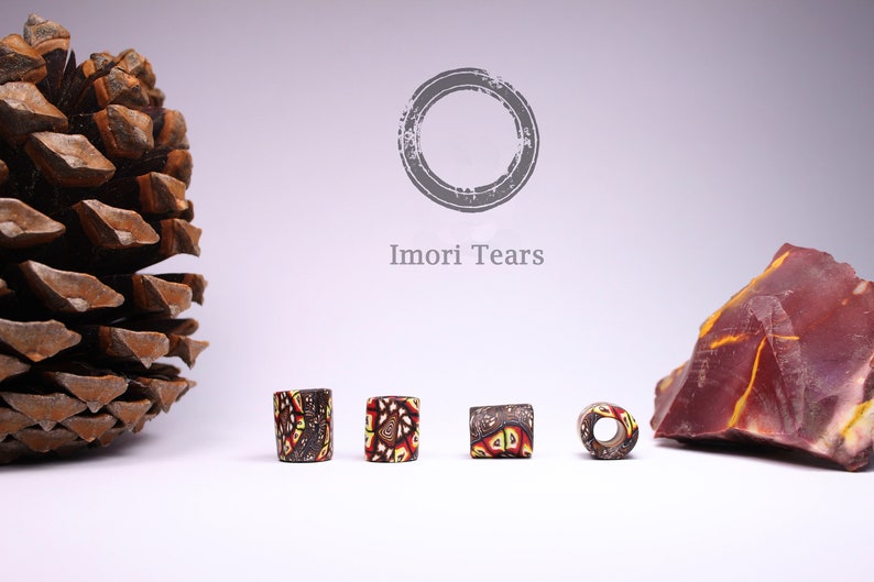 May include: Four colorful glass beads with a geometric pattern in shades of brown, red, and yellow. The beads are arranged in a row on a white surface. The text "I mori Tears" is visible in the background.