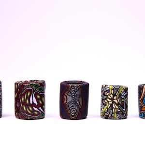 May include: Five colorful cylindrical ceramic beads with intricate patterns. The beads are arranged in a row on a white surface. The patterns include swirls, geometric shapes, and floral designs. The beads are a mix of brown, white, red, blue, green, and yellow.