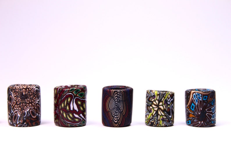 May include: Five colorful cylindrical ceramic beads with intricate patterns. The beads are arranged in a row on a white surface. The patterns include swirls, geometric shapes, and floral designs. The beads are a mix of brown, white, red, blue, green, and yellow.