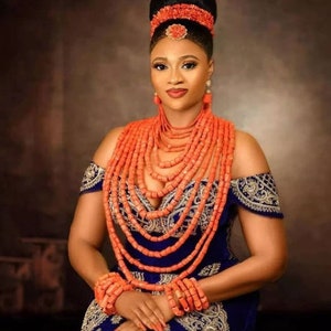 Puede incluir: Una mujer con un vestido azul y dorado con un collar tradicional de cuentas de coral y pulseras a juego. Las cuentas de coral son de un color naranja vibrante y están dispuestas en varias hileras. La mujer lleva el pelo recogido en un moño alto con un tocado de cuentas de coral.