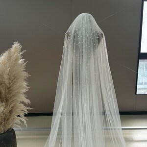 May include: A white tulle wedding veil with a delicate pearl embellishment. The veil is long and flowing, with a soft, romantic look.