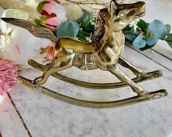 May include: A vintage brass rocking horse figurine. The horse is detailed with a saddle and flowing mane and tail. The figurine is approximately 6 inches long and sits on curved rockers.
