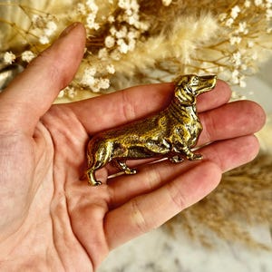 Könnte beinhalten: Eine kleine, goldfarbene Dackel-Figur. Der Hund ist in einer stehenden Position dargestellt, mit leicht erhobenem Kopf und Schwanz. Die Figur wird in einer Hand gehalten, mit einem verschwommenen Hintergrund aus getrockneten Blumen.
