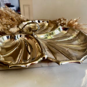 Puede incluir: Una fuente para servir de color plateado con tres secciones y un diseño en forma de hoja. La fuente tiene un asa y un borde festoneado. La fuente es de metal y probablemente se utiliza para servir aperitivos o bocadillos.