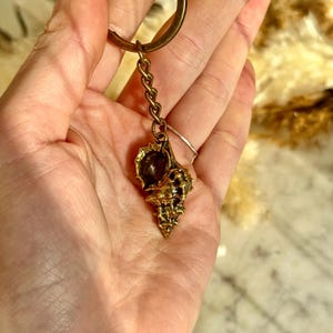 Könnte beinhalten: Ein goldfarbener Schlüsselanhänger mit einem detaillierten Muschel-Anhänger. Der Anhänger ist an einer Kette und einem runden Schlüsselring befestigt. Die Muschel hat aufwendige Texturen und ein naturalistisches Design.