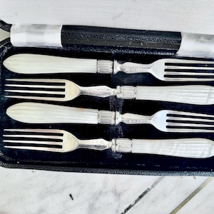 May include: Four antique forks with mother-of-pearl handles, presented in a black case with a velvet lining. The forks have three tines and silver-coloured metal. The handles are a light cream colour.