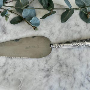 Puede incluir: Servidor de pastel plateado con un mango decorativo. El servidor tiene un borde dentado y el texto "Pie Server" es visible en la hoja. El mango presenta diseños florales y de volutas.