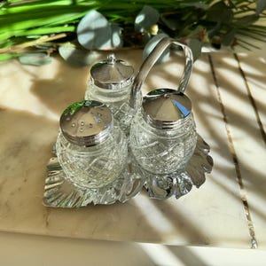 May include: A vintage salt and pepper set with a sugar bowl, featuring clear, cut-glass containers with silver-toned lids and a handle. The set sits on a decorative silver-toned tray, with a marble-like surface in the background.