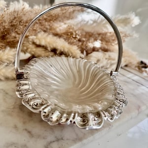 Puede incluir: Una cesta ovalada de cristal transparente con asa plateada y bordes ornamentados y detallados. El interior de cristal tiene un diseño en espiral. El asa es arqueada y la cesta se apoya sobre una superficie de mármol.