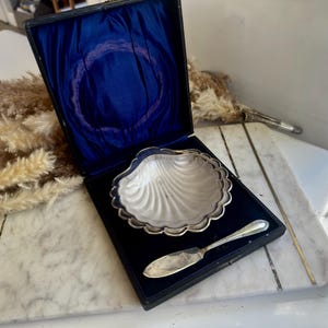May include: A vintage silver-plated seashell-shaped dish with a scalloped edge, presented in a black box with a blue velvet lining. A small silver knife rests beside the dish, suggesting a serving set.