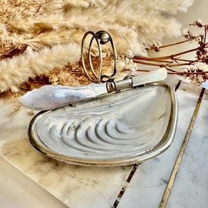 Pode incluir: Uma manteigueira vintage prateada em forma de concha, com uma faca de manteiga a condizer e um porta-manteiga prateado. O prato e a faca repousam sobre uma superfície com padrão de mármore, com flores secas no fundo.