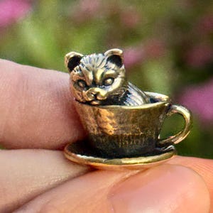 Puede incluir: Una pequeña figura de gato de color bronce sentada dentro de una taza de té. El gato tiene detalles finos, y la taza de té incluye un platillo y un asa. El diseño general es en miniatura y detallado, con una estética vintage.