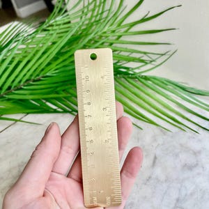 Op de afbeelding: Een messing liniaal met centimeter markeringen, gehouden tegen een achtergrond van groene palmbladeren. De liniaal is ongeveer 10 cm lang en heeft een gat aan de bovenkant om op te hangen.