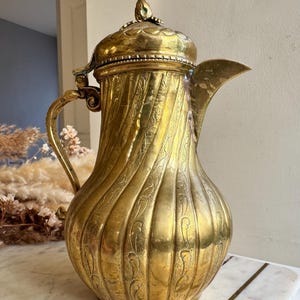 May include: A vintage brass pitcher with a decorative lid and handle. The pitcher has a spiral design with etched floral patterns. The spout is curved, and the overall color is a shiny gold. The pitcher is likely used for serving beverages.