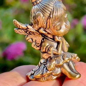 Könnte beinhalten: Kleine, detaillierte Messingfigur eines sitzenden Mädchens mit Dutt-Frisur, das ein Blatt hält. Die Figur hat eine Vintage-Ästhetik mit aufwendigen Details in Haar und Kleidung. Das Mädchen trägt ein Kleid mit Rüschenrock.