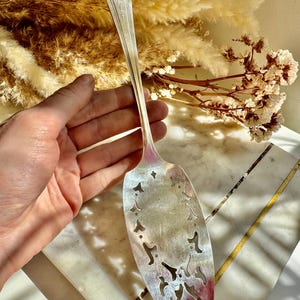 Puede incluir: Un servidor de pastel de plata vintage con un diseño calado decorativo. El servidor tiene un mango largo y una hoja ovalada ancha con patrones florales recortados. La hoja tiene un ligero tono rojizo cerca del borde, y el mango tiene un diseño floral grabado.