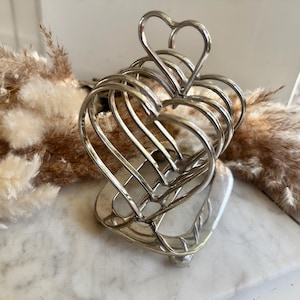May include: A silver-colored heart-shaped napkin holder. The holder is made of curved metal wires forming multiple heart shapes. The base is a solid heart shape with a small round foot. The top features a single heart.