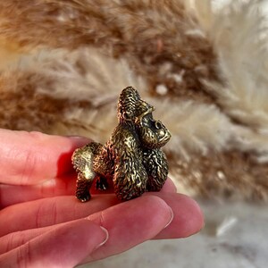 May include: A small, bronze gorilla figurine with detailed fur and facial features. The gorilla is in a standing pose. The figurine is held in a hand, with a blurred background of dried plants.