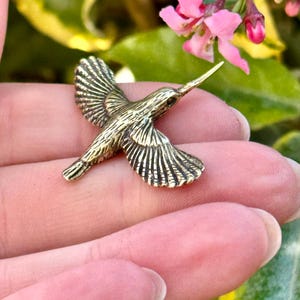 Puede incluir: Un colgante detallado de colibrí dorado con las alas extendidas. Las plumas del pájaro están intrincadamente detalladas. El fondo incluye flores rosas y follaje verde, creando un entorno natural. El colgante se sostiene en una mano.