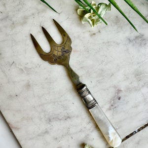 May include: An antique serving fork with three tines. The fork's head is decorated with floral engravings. The handle is made of mother-of-pearl and metal. The fork is approximately 18 cm long.