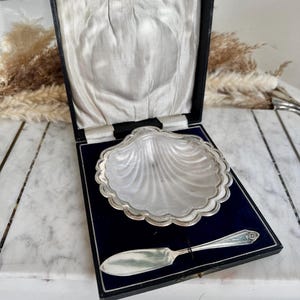 Puede incluir: Un plato de mantequilla vintage plateado en forma de concha de vieira, presentado en una caja negra con interior de terciopelo azul. Incluye un cuchillo de mantequilla a juego. El plato mide aproximadamente 13 cm de diámetro.