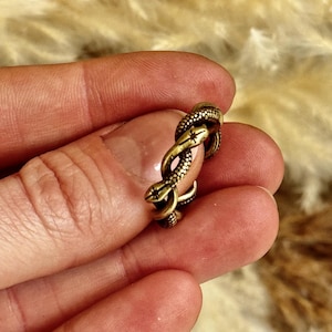 May include: A bronze ring with a snake design. The snake is coiled around the ring, with detailed scales and a textured appearance. The ring is held in a hand against a blurred background.