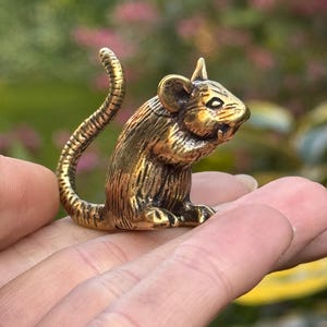 Puede incluir: Pequeña y detallada figura de rata de latón. La rata está en posición sentada con la cola curvada hacia arriba. La figura tiene una superficie texturizada y se sostiene en una mano sobre un fondo borroso verde y rosa.