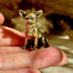 May include: A small, antique-style brass figurine of a chihuahua dog in a seated position. The metal sculpture features detailed textures representing fur, pointed ears, and alert eyes, with a dark, weathered patina finish across the surface.