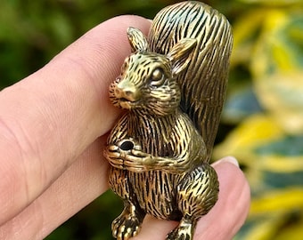 May include: A detailed brass squirrel figurine. The squirrel is depicted in a seated position, holding something in its paws. The figurine has a textured surface and a bushy tail. The background is blurred, with green and yellow foliage.
