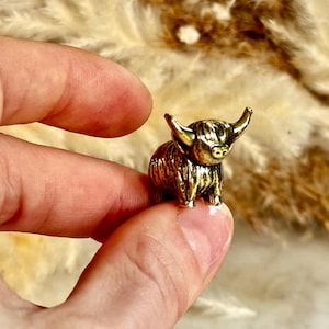 Op de afbeelding: Een kleine, gedetailleerde goudkleurige Highland-koeienfiguur. De miniatuursculptuur heeft opvallende hoorns en een getextureerde vacht, vastgehouden tussen vingers tegen een vage, neutrale achtergrond. De koe is een charmant decoratief item.
