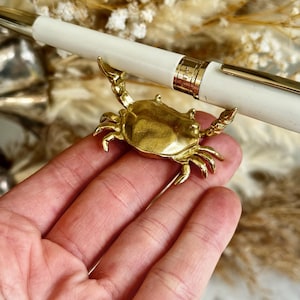 May include: A gold-colored crab pen holder supporting a white pen with gold accents. The crab is detailed with claws and a textured shell. The pen has a gold band near the tip and a gold clip. The background features dried flowers.
